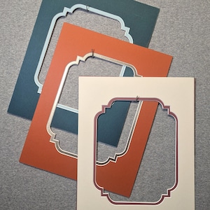 May include: Three rectangular picture frame mats layered over each other. The top mat is cream with a dark red inner border, the middle is burnt orange with a gold inner border, and the bottom is dark teal with a light blue inner border.