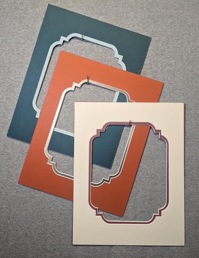 May include: Three rectangular picture frame mats layered over each other. The top mat is cream with a dark red inner border, the middle is burnt orange with a gold inner border, and the bottom is dark teal with a light blue inner border.
