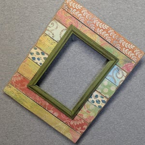 May include: A decorative picture frame with a green inner frame and a larger outer frame. The outer frame has a colorful, distressed design with floral and geometric patterns in shades of pink, orange, yellow, and blue.