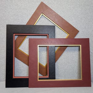 Può includere: Tre cornici rettangolari sovrapposte. La cornice posteriore è marrone con un bordo interno dorato, quella centrale è nera con un bordo interno color corallo e la cornice anteriore è rosso scuro con un bordo interno dorato.