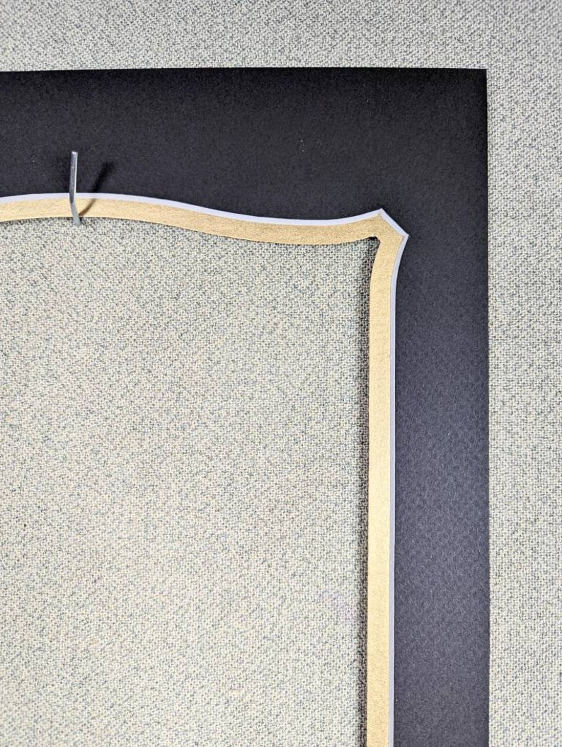 May include: Close-up of a corner of a picture frame with a decorative, scalloped, gold-colored mat. The mat is set against a black background.