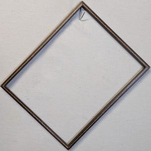 May include: A dark brown picture frame with a decorative gold-colored trim. The frame is empty and has a metal hook at the top for hanging. The frame is square-shaped and angled, showcasing a minimalist design.