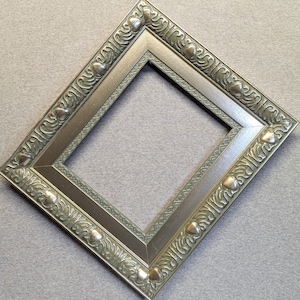 May include: A silver picture frame with an ornate, green, leaf-like design. The frame has a raised edge and a beveled mat.
