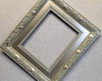 May include: A silver picture frame with an ornate, green, leaf-like design. The frame has a raised edge and a beveled mat.