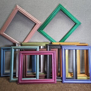 May include: A collection of eight picture frames in various colors and sizes, including pink, green, brown, blue, and silver. The frames are arranged in a staggered pattern, with the largest frame at the top and the smallest frames at the bottom.