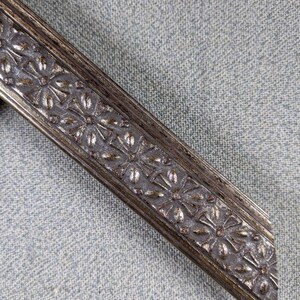 May include: Close-up of a silver picture frame with a decorative floral design. The frame is made of metal and has a distressed finish.