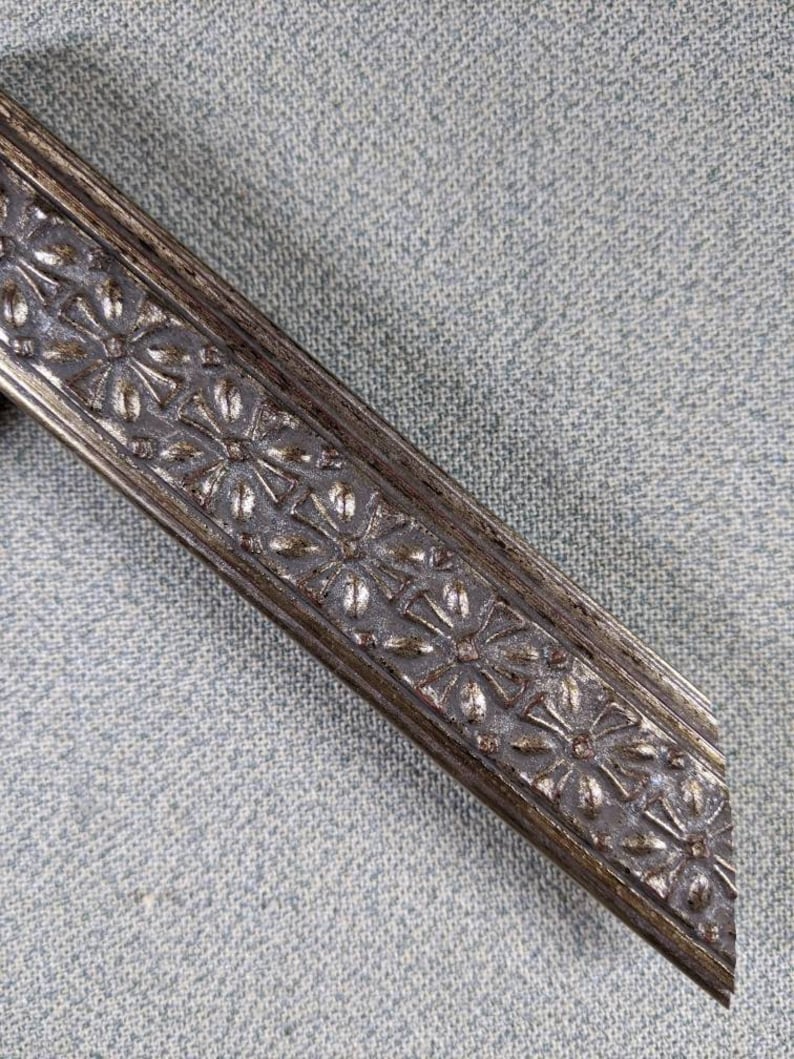May include: Close-up of a silver picture frame with a decorative floral design. The frame is made of metal and has a distressed finish.