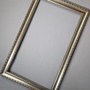 May include: A rectangular silver picture frame with a decorative rope-like border. The frame has a slightly distressed finish, giving it a vintage appearance. The frame is empty, ready for a photo or artwork.