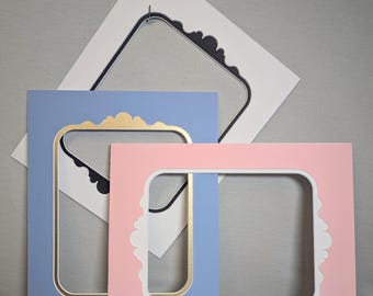 May include: Three decorative picture frame mats in pastel colors. A light blue mat with a gold inner border, a pink mat with a white inner border, and a white mat with a black inner border. The mats have decorative, scalloped edges.