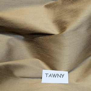 Pu&ograve; includere: Primo piano di un tessuto piegato di colore fulvo. Il tessuto ha una lucentezza sottile e una trama visibile, con aree chiare e scure che creano profondit&agrave;. Un'etichetta bianca con la parola "TAWNY" si trova nell'angolo in basso a destra.
