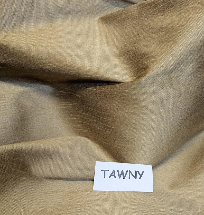 Pu&ograve; includere: Primo piano di un tessuto piegato di colore fulvo. Il tessuto ha una lucentezza sottile e una trama visibile, con aree chiare e scure che creano profondit&agrave;. Un'etichetta bianca con la parola "TAWNY" si trova nell'angolo in basso a destra.