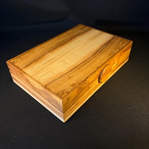 May include: A rectangular wooden box featuring a prominent natural grain pattern with light and dark brown streaks. The box has a smooth, flat lid and a small, circular wooden knob on the front side for opening, showcasing the organic wood texture.