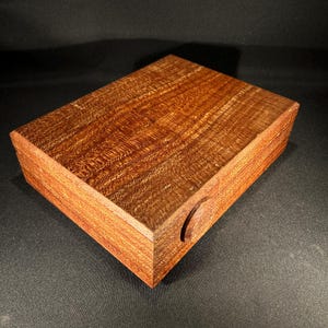 May include: A rectangular wooden box with a rich, reddish-brown hue. The box features a lid and a circular indentation on one side for easy opening. The wood grain is prominent, adding texture and visual interest to the handcrafted item.