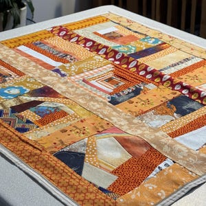 May include: A rectangular patchwork quilt featuring a variety of fabric patterns in shades of orange, yellow, brown, and cream. The design consists of numerous small, rectangular fabric strips sewn together in a log cabin style arrangement.