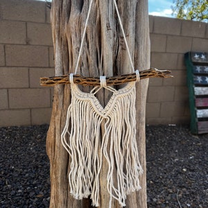 May include: A white macrame wall hanging with a wooden branch at the top. The macrame is hanging from a tree trunk.
