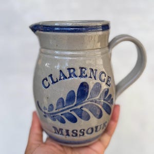 May include: A gray ceramic pitcher with a blue rim and handle. The pitcher features the text "CLARENCE MISSOURI" and a blue leaf design. The pitcher has a spout for pouring.