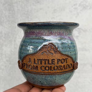 May include: A small, handcrafted ceramic pot with a blue, purple, and brown glaze. The pot features a brown plaque with the text "A Little Pot From Colorado" and a mountain design. The pot is held by a hand.