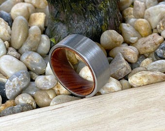 May include: A ring featuring a brushed grey metal exterior band and a smooth reddish-brown wood interior lining. The band has a flat profile and a wide construction, showcasing a contrast between the metallic outer surface and the natural wood grain.