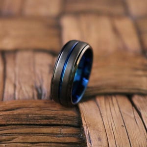 May include: A matte black ring featuring a thin, polished blue groove running horizontally around the center of the band. The interior of the ring is also finished in a polished blue tone, creating a contrast with the dark exterior surface.