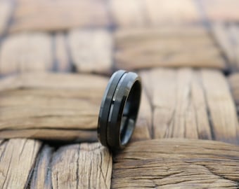 Op de afbeelding: Een zwarte ring met een gladde, matte afwerking en een dunne, gepolijste zilverkleurige groef die horizontaal rond het midden van de band loopt. De ring heeft een plat profiel met afgeschuinde randen voor een strak en eenvoudig ontwerp.