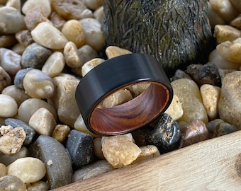 May include: A ring featuring a matte black outer band with a smooth, brushed finish and a polished reddish-brown wood grain interior lining. The band has a flat profile and a wide construction, showcasing a contrast between the dark metal and wood.