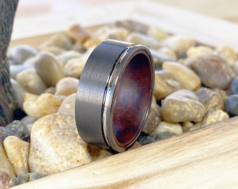 May include: A ring featuring a brushed dark gray metal exterior band with a polished silver-tone edge and a smooth, deep reddish-brown wood grain inlay on the interior surface, showcasing a distinct contrast between the metal and wood materials.