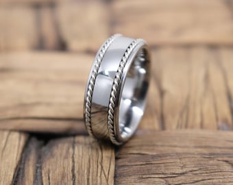 May include: A silver-toned metal ring featuring a smooth, polished center band flanked by two detailed rope-style textured edges. The ring has a rounded profile and a reflective surface that catches light, showcasing a clean and metallic finish.