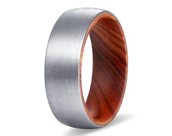 May include: A ring featuring a brushed silver-toned metal exterior and a smooth, reddish-brown wood grain interior. The band has a rounded profile with a matte finish on the outer surface, contrasting with the polished, warm-toned wooden lining.