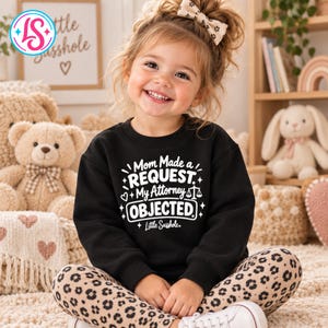 May include: A black long-sleeve sweatshirt featuring white text that reads Mom Made a REQUEST My Attorney OBJECTED with a scale of justice icon and the text Little Sasshole. The sweatshirt is paired with tan and black leopard print leggings.