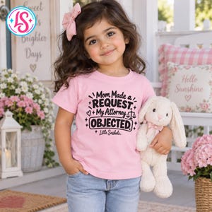 May include: A light pink short-sleeved t-shirt featuring black text that reads Mom Made a REQUEST My Attorney OBJECTED with a small scale of justice icon and the text Little Sasshole. The shirt is paired with light blue denim jeans.