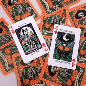 Può includere: Carte da gioco a tema Halloween con illustrazioni di scheletri e zucche. Una carta mostra uno scheletro di gatto, l'altra una farfalla con un teschio. Le carte sono circondate da altre carte con disegni simili.