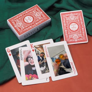 May include: A personalised deck of playing cards with a red and white design. The card faces feature personal photos. The card box and the backs of the cards have the names "Gerret & Kacey" and the date "05.06.25".