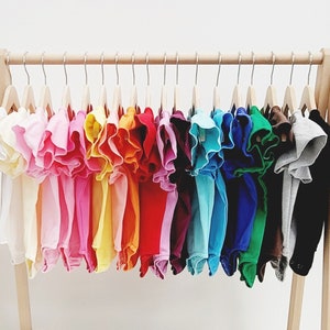 May include: A wooden rack with a variety of colorful ruffle-trimmed shirts hanging on hangers. The shirts are arranged in a rainbow pattern, with the colors ranging from white to dark grey.