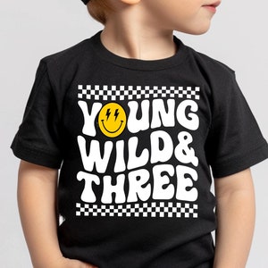 May include: A black t-shirt with a white graphic that says "Young Wild & Three" with a yellow smiley face with lightning bolts for eyes. The shirt is being worn by a child wearing a black baseball cap.
