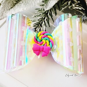 May include: A holographic iridescent bow with a white heart pattern and a pink and yellow swirl lollipop with a pink bow on top.
