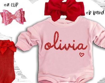 May include: Pink baby onesie with the name "Olivia" in red glitter lettering and a small red glitter heart. The onesie is paired with a red glitter hair clip, a red knit headband, and red knit tights.