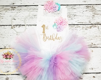 Peut inclure: Un tutu blanc avec des couches de tulle rose, bleu et violet. Un body blanc avec un texte pailleté doré qui dit "1er anniversaire" et un embellissement floral est sur le dessus du tutu.