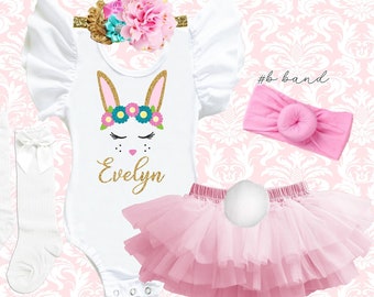 May include: A white bunny-themed outfit for a baby or toddler, including a bodysuit, tutu, headband, and socks. The bodysuit features a bunny face design with the name "Evelyn" in gold glitter. The tutu is pink, and the headband has floral accents.