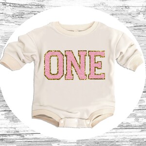 May include: A white long-sleeved onesie with a pink and gold glitter "ONE" graphic.
