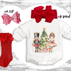 May include: White long-sleeved onesie with a watercolor illustration of a nutcracker and a ballerina standing in front of a Christmas tree with presents. The onesie is on a wooden background. There is a red headband with a bow, a red pair of tights with a bow, and a pink glitter hair clip.
