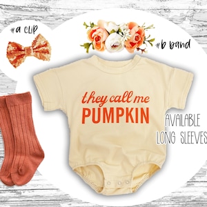 May include: A beige baby onesie with the text "they call me PUMPKIN" in orange. The onesie is paired with a pair of orange ribbed knee-high socks, a sparkly orange bow, and a flower crown.