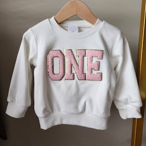 Peut inclure: Sweat-shirt blanc avec une applique "UN" rose et or sur le devant.