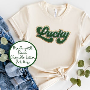 May include: Cream-colored t-shirt with the word "Lucky" in a green chenille letter patch with gold glitter trim. The shirt is folded, with a denim jacket and greenery in the background. The text "Made with Real Chenille Letter Patches!" is in a green circle.
