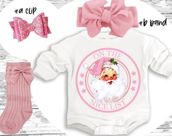 May include: A white baby onesie with a pink circle graphic featuring a cartoon Santa Claus with the text "On the Nice List". The onesie is paired with a pink headband, pink socks, and a pink glitter hair bow.