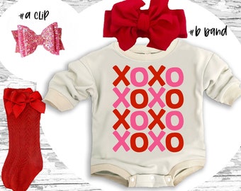 Könnte beinhalten: Ein weißer Baby-Strampler mit dem Text "XOXO", der viermal in Rot und Pink wiederholt wird. Der Strampler wird mit einem roten Stirnband, roten Socken mit Schleife und einer pinkfarbenen Glitzer-Haarklammer kombiniert.