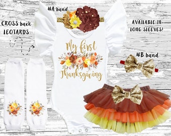 May include: A white bodysuit with the text "My first Thanksgiving" in gold glitter lettering and a floral wreath design. The bodysuit is paired with a brown tulle skirt with yellow, orange, and red layers. The outfit includes two leg warmers with a floral design and two headbands, one with a brown flower and the other with a gold bow.