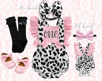 May include: A baby outfit set featuring a cow print romper with pink accents and the word "one". The set includes a matching headband, socks, shoes, and a second headband. The romper has ruffles and bows.