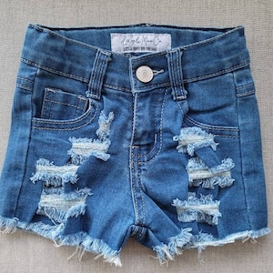 May include: Distressed denim shorts with frayed edges. The shorts have a light blue wash and are made of a soft, comfortable fabric. The brand name "Little Miss Co." is printed on the back waistband.