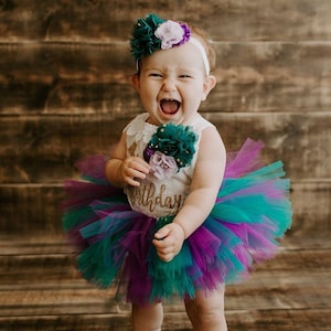 May include: A baby girl wearing a white onesie with gold glitter text that says "1st Birthday" and a green and purple tulle tutu. She is wearing a headband with green and purple flowers. The baby is smiling and laughing.