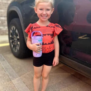 May include: A young girl wearing a red and black patterned top and black shorts smiles while holding a purple water bottle. The water bottle has a handle and a design on it. The girl is standing next to a black vehicle.